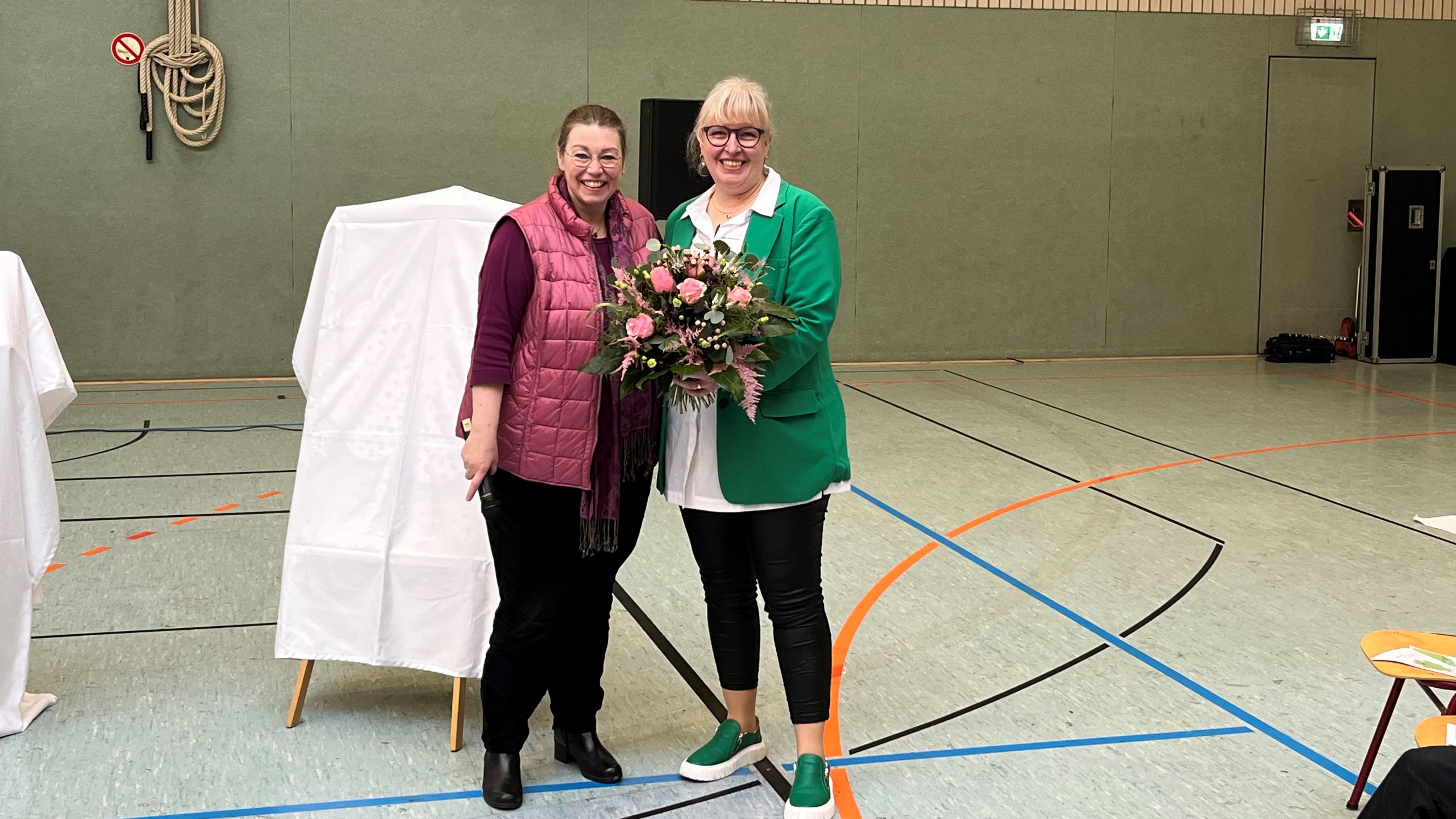 Die Erste Stadträtin Sandra Sollmann und Anja Heckmann-Hollmann stehen in einer Turnhalle. Heckmann-Hollmann wird ein Blumenstraß überreicht