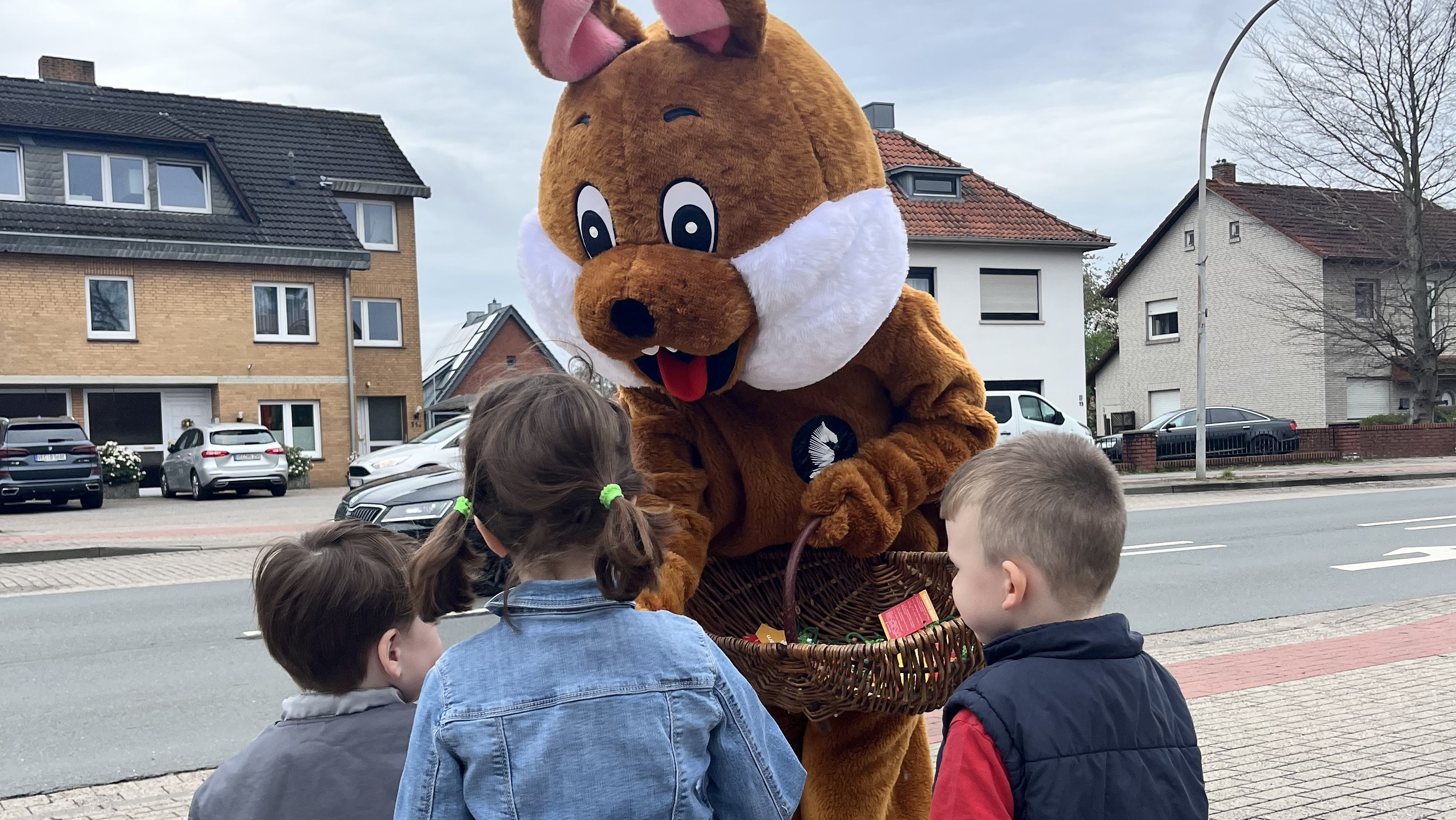 Ein mannshoher Osterhase steht vor 3 Kindern mit einem Korb und verteilt etwas an sie.